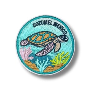 May include: A circular embroidered patch featuring a sea turtle swimming in turquoise water with coral reefs at the bottom. The text COZUMEL MEXICO is stitched in dark blue along the top edge. The turtle has a brown shell and pinkish fins.