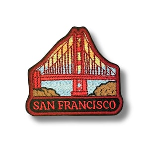 Peut inclure: Un écusson brodé représentant le Golden Gate Bridge avec des fils rouges, jaunes et bleu clair sur un fond sombre. Le pont est flanqué de terrains rocheux bruns à la base. Le texte SAN FRANCISCO est brodé en rouge sur la partie inférieure.