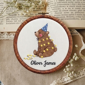 Può includere: Un telaio da ricamo circolare in legno contenente tessuto bianco con un orso marrone ricamato a mano che indossa un cappellino da festa blu a pois. L'orso è avvolto da una catena di luci gialle e il nome Oliver James è ricamato in nero.