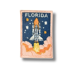 May include: Embroidered patch featuring a space shuttle launching into space. The patch has a blue background with the word "FLORIDA" at the top. The shuttle is white and gray with orange flames. The patch is bordered in a light peach color.