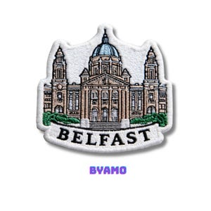 May include: Embroidered patch featuring Belfast City Hall. The patch is white with a detailed illustration of the building in shades of brown, beige, and blue. The word "BELFAST" is embroidered in black, with "BYAMO" in purple below.