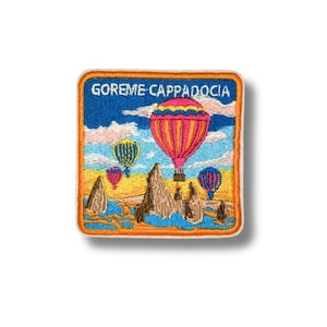 May include: Embroidered patch featuring a vibrant scene of hot air balloons over a unique landscape. The patch has a blue sky with the text "GOREME CAPPADOCIA" at the top. The patch is bordered with an orange frame.