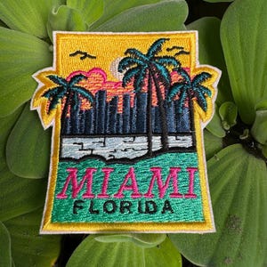 May include: A yellow, green and pink embroidered patch featuring a sunset scene, palm trees and the text "MIAMI FLORIDA".