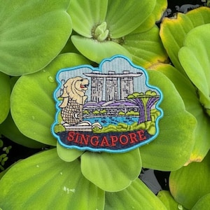 May include: Embroidered patch depicting the Merlion statue and the Marina Bay Sands in Singapore. The patch has a blue border and the word "SINGAPORE" in red. The patch is set against a background of green aquatic plants.