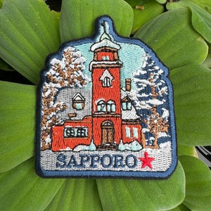 May include: A red embroidered patch with a white building and snow-covered trees. The text "SAPPORO" is in blue with a red star.