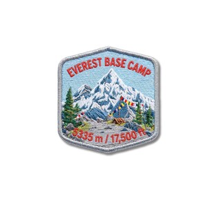 Può includere: Una toppa ricamata esagonale raffigurante un paesaggio montano innevato con pini verdi e bandiere di preghiera colorate. Il testo EVEREST BASE CAMP è in rosso in alto, e 5335 m / 17.500 ft è in rosso in basso, su uno sfondo azzurro.