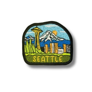 May include: An embroidered patch with a black border featuring a colorful scene of the Seattle skyline, including the Space Needle, a snow-capped mountain, and a yellow sun against a blue sky. The word SEATTLE is stitched in yellow at the bottom.