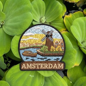 May include: Embroidered patch featuring a Dutch landscape with a windmill, boat, and houses. The patch has a circular design with the word "AMSTERDAM" in a rectangular banner at the bottom. The patch is surrounded by green aquatic plants.
