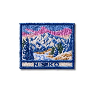 May include: A rectangular embroidered patch featuring a snowy mountain landscape with dark green pine trees and a pink and blue sky. The word NISEKO is stitched in blue capital letters on a white horizontal band at the bottom of the patch.