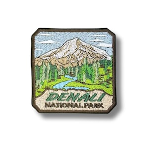 May include: A square embroidered patch featuring a snow-capped mountain peak above a green forest and a winding blue river. The bottom section contains the text DENALI NATIONAL PARK in dark green and black lettering with a dark brown border.