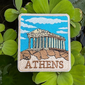 May include: Square embroidered patch depicting the Parthenon in Athens, Greece, with a blue sky and white clouds. The patch has a light blue border and the word "ATHENS" in tan. The patch is surrounded by green plants, floating in water.