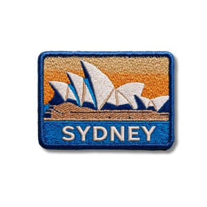 May include: A rectangular embroidered patch featuring the Sydney Opera House in white against a golden orange sky. The base shows blue water and a brown shoreline. The word SYDNEY is stitched in white capital letters across the bottom blue section.