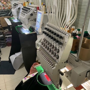 Puede incluir: Una m&aacute;quina de bordar industrial de m&uacute;ltiples agujas con estructura blanca, numerosos carretes de hilo y una pantalla de control digital. La m&aacute;quina cuenta con un bastidor circular verde que sujeta una tela negra con un elemento rojo.