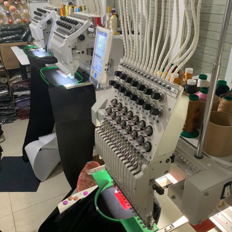Puede incluir: Una m&aacute;quina de bordar industrial de m&uacute;ltiples agujas con estructura blanca, numerosos carretes de hilo y una pantalla de control digital. La m&aacute;quina cuenta con un bastidor circular verde que sujeta una tela negra con un elemento rojo.