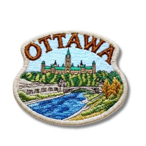 Puede incluir: Parche bordado con la palabra "OTTAWA" en marrón sobre una representación detallada de los edificios del Parlamento de Ottawa, un río y un puente. El parche tiene un borde beige claro y un fondo de cielo azul.
