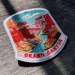 May include: A shield-shaped embroidered patch featuring a landscape of the Grand Canyon with red rock formations, a winding blue river, and a light blue sky. The text GRAND CANYON is stitched in red letters along the bottom white border.