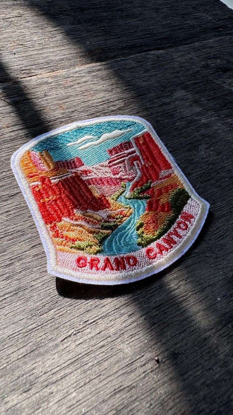 May include: A shield-shaped embroidered patch featuring a landscape of the Grand Canyon with red rock formations, a winding blue river, and a light blue sky. The text GRAND CANYON is stitched in red letters along the bottom white border.