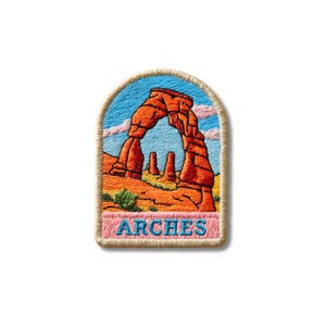 May include: An embroidered patch with an arched top featuring a landscape of orange and red rock formations under a blue sky with pink clouds. The word ARCHES is stitched in blue capital letters on a light pink rectangular background at the base.
