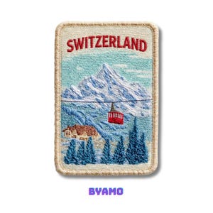 Op de afbeelding: Geborduurde patch met een Zwitsers berglandschap met een rode kabelbaan, besneeuwde bergtoppen en het woord "SWITZERLAND" in het rood. De patch heeft een crèmekleurige rand en het woord "BYAMO" in paars.