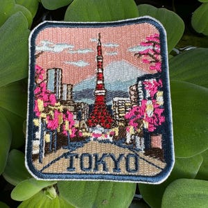 May include: Embroidered patch depicting Tokyo, featuring the Tokyo Tower, cherry blossoms, and the word "TOKYO". The square patch has a navy border and a pink sky. The design includes buildings and a mountain, with the overall theme of a city scene.