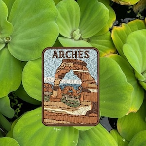 May include: Embroidered patch featuring the word "ARCHES" above an image of a sandstone arch in a desert landscape. The patch has a brown border and is set against a background of green aquatic plants.
