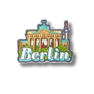 Puede incluir: Un parche bordado que muestra la Puerta de Brandeburgo en hilos beige y marrón con follaje verde en la base y una torre alta a la derecha. La palabra Berlin está bordada en letras blancas grandes con un contorno azul claro en la parte inferior.