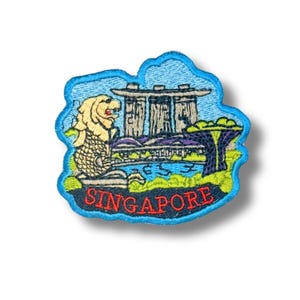 Op de afbeelding: Een geborduurde patch met het Merlion-standbeeld en het Marina Bay Sands-hotel in Singapore. Het ontwerp bevat groen gebladerte, een blauwe lucht en het woord SINGAPORE in rood garen aan de onderkant, omrand door een lichtblauwe rand.