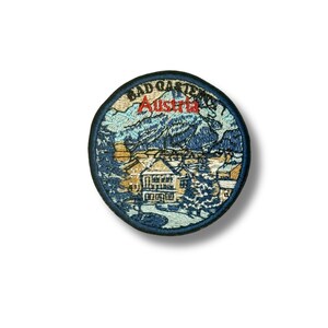 May include: Circular embroidered patch depicting a mountain scene with buildings. The text "BAD GASTEIN Austria" is arched around the top in a dark blue font, with "Austria" in red.