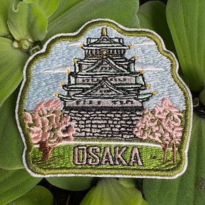 以下が含まれることがあります： 緑と白の大阪城が刺繍されたパッチ。両側にピンクの桜の花が咲いています。パッチは緑色の背景に白い文字で「OSAKA」と書いてあります。