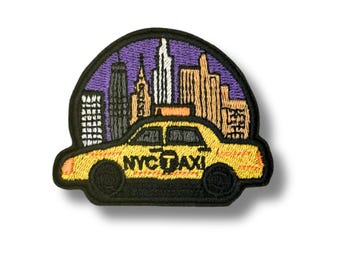 May include: An embroidered patch featuring a yellow taxi cab with the text NYC TAXI on the side. The background shows a stylized city skyline in shades of purple, tan, and white, all enclosed within a black border with a rounded top edge.