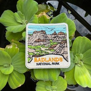 May include: Embroidered patch depicting the Badlands National Park landscape. The square patch shows a light blue sky, brown rock formations, and green vegetation. The text "BADLANDS NATIONAL PARK" is in orange and white.