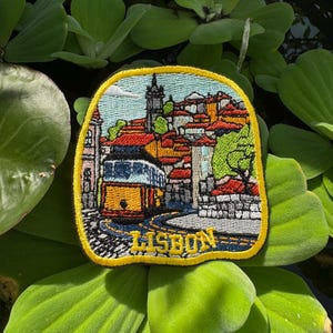 Può includere: Una toppa ricamata gialla con un'immagine colorata di un tram a Lisbona, Portogallo. La toppa ha un contorno nero e un bordo giallo con la parola "LISBONA" ricamata in giallo.