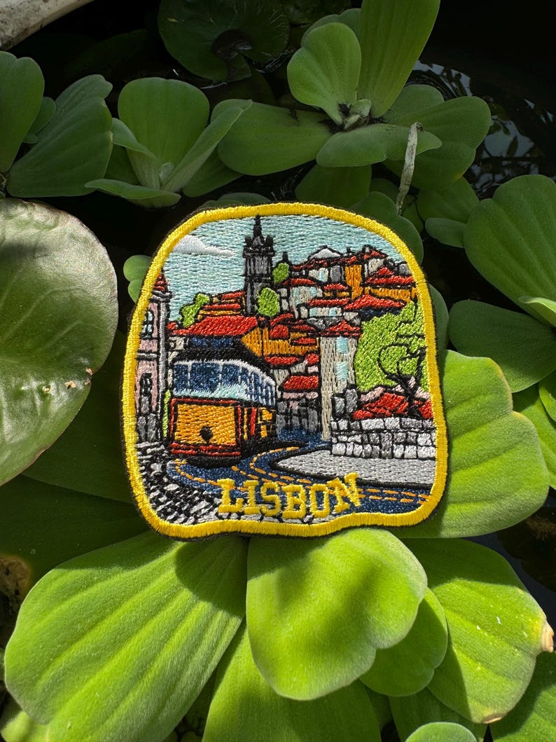 May include: Embroidered patch featuring a colorful depiction of Lisbon, Portugal. The design includes a yellow tram, red-tiled roofs, and the word "LISBON" in yellow. The patch has a yellow border and is set against a background of green leaves.