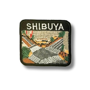 Puede incluir: Un parche bordado cuadrado con fondo negro y el texto blanco SHIBUYA en la parte superior. El diseño muestra una vista aérea de una intersección urbana con pasos de cebra blancos, carreteras grises, árboles verdes y edificios color canela.