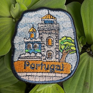 Könnte beinhalten: Gestickter Aufnäher mit einem grauen Schloss mit blauem Dach und einem gelben Turm. Das Schloss ist von einem grünen Baum und einem blauen Himmel umgeben. Der Text "Portugal" ist in Orange auf einem braunen Banner gestickt.