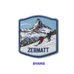 May include: Embroidered patch featuring a snowy mountain scene with two skiers. The patch has a blue border and the word "ZERMATT" is displayed in white. The word "BYAMO" is displayed in purple.