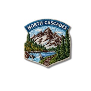 May include: An embroidered patch featuring a mountain landscape with snow-capped peaks, a blue lake, and green pine trees. The text NORTH CASCADES is stitched in white on a blue arched banner at the top of the patch, which has a light border.