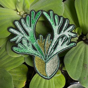 May include: Green and gold embroidered patch of a stylised plant with a rounded base and multiple branching stems.