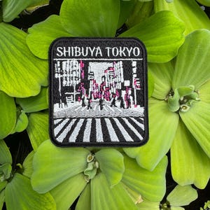 May include: A rectangular embroidered patch featuring a black and white scene of the Shibuya Crossing in Tokyo. The text SHIBUYA TOKYO is stitched in white at the top. The design includes a crosswalk, city buildings, and figures in black and white.