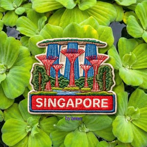 Puede incluir: Parche bordado con el horizonte de Singapur, incluyendo el Supertree Grove y el hotel Marina Bay Sands. El parche tiene la palabra "SINGAPORE" en rojo sobre un fondo azul. El parche está rodeado de plantas verdes.