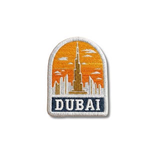 Può includere: Una toppa ricamata che raffigura lo skyline di Dubai con il Burj Khalifa al centro su un cielo arancione al tramonto. La toppa ha una parte superiore ad arco e una base rettangolare blu scuro con il testo bianco DUBAI stampato sopra.