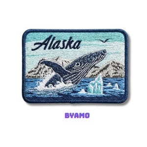 Puede incluir: Parche bordado que representa una ballena saltando del mar, con la palabra "Alaska" encima. El parche tiene un borde azul e incluye montañas, icebergs y un pájaro. El texto "BYAMO" está en la parte inferior.