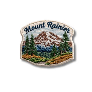 May include: An embroidered patch featuring a snow-capped mountain landscape with green pine trees and golden-brown foreground foliage. The text Mount Rainier is stitched in dark blue thread across the top of the curved, cream-bordered patch.
