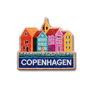 Può includere: Una toppa ricamata che raffigura una fila di case colorate in arancione, turchese, rosa e giallo con una guglia centrale. Sotto gli edifici si trova uno stendardo rettangolare blu scuro con il testo bianco COPENHAGEN e un bordo in pietra.