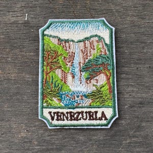 Könnte beinhalten: Gestickter Aufnäher mit einer Wasserfallszene mit üppigem grünen Laub, braunen Klippen und einem blauen Wasserbecken. Der Aufnäher hat einen weißen Rand und das Wort "VENEZUELA" in braunen Buchstaben.
