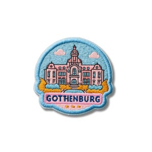 May include: A circular embroidered patch featuring a light pink building with dark blue outlines against a light blue sky with two small pink clouds. The word GOTHENBURG is stitched in dark blue letters on a pink banner at the bottom.