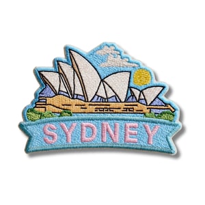 May include: An embroidered patch featuring the Sydney Opera House with white sails, a yellow sun, and light blue clouds against a blue sky. Below the building, a light blue banner displays the word SYDNEY in large, light pink capital letters.