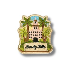 May include: Embroidered patch featuring a pink building with a tower, palm trees, and the text "Beverly Hills." The patch has a beige border and a blue sky background. The design is detailed and colorful, suitable for clothing or accessories.