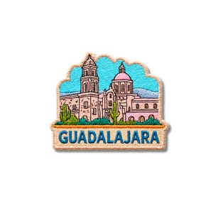Pode incluir: Um emblema bordado apresentando uma catedral rosa com uma torre alta e cúpula contra um céu azul brilhante. Cactos verdes estão posicionados em primeiro plano. A palavra GUADALAJARA está bordada em letras maiúsculas azuis numa borda bege.