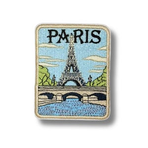 May include: A rectangular embroidered patch featuring the Eiffel Tower in the center with the word PARIS in black text above it. The design includes a light blue sky with clouds, green trees, a stone bridge, and blue water in the foreground.